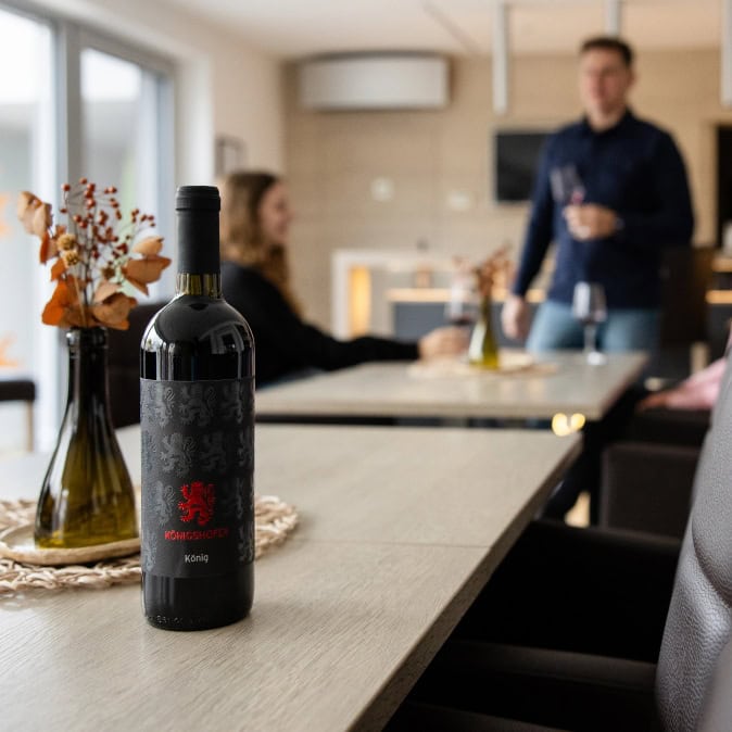 Rotweinflasche König steht auf Tisch, im Hintergrund verschwommene Personen.
